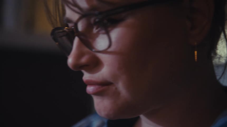 The video transitions from a closeup of dynamic financial graphs on a screen to a profile shot of a woman with glasses who is intently focused on her work in a dimly lit professional setting
