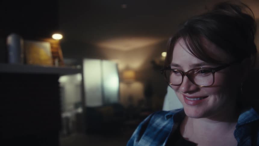 Working late at her desk a woman studies data on two screens then smiles with a sense of accomplishment and takes off her glasses