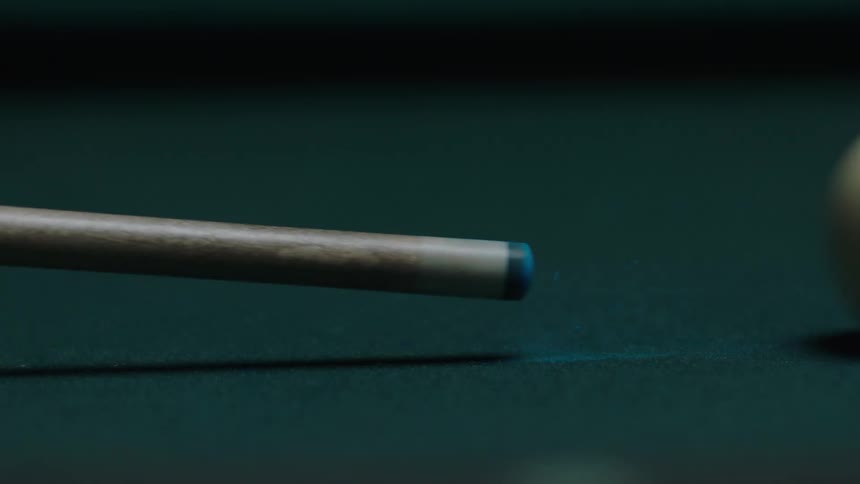A closeup shot of a white cue ball resting on a green pool table felt is interrupted when a cue stick showing its blue chalked tip moves into the frame anticipating a shot