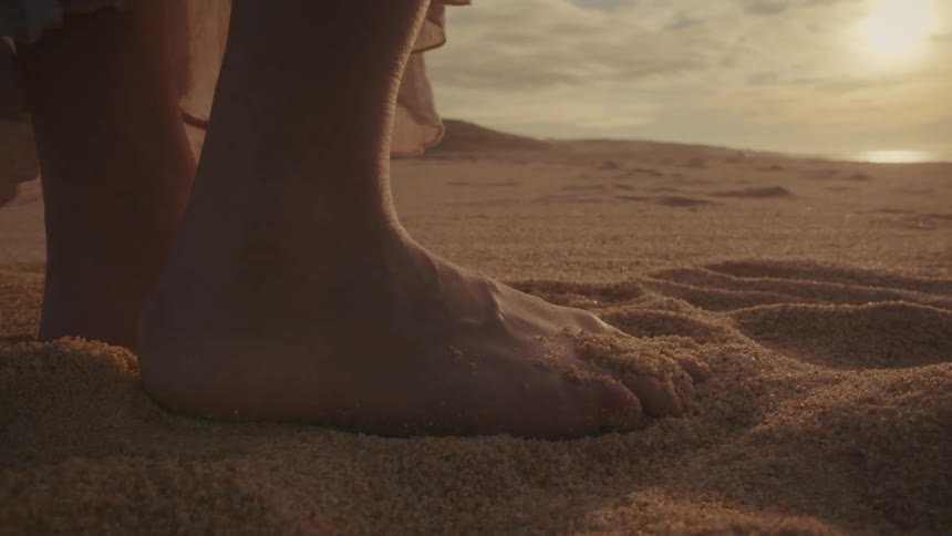 Barefoot woman stands on the beach as her long dress moves in the breeze