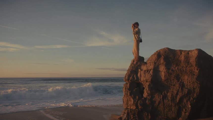A young male photographer taking photos of nature while standing on a cliff at the beach at sunset