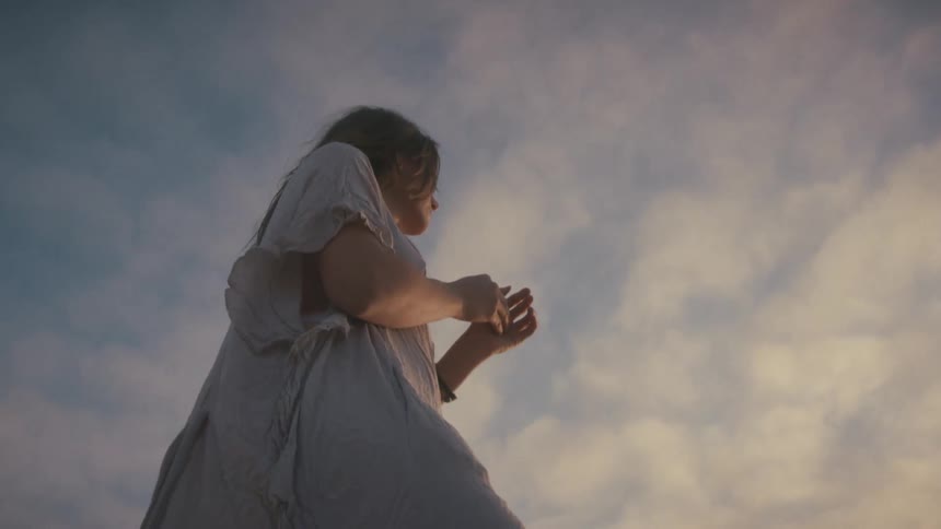 A female is wearing a lightcolored flowing dress against a dramatic sky filled with scattered clouds with warm light highlighting the scene from behind