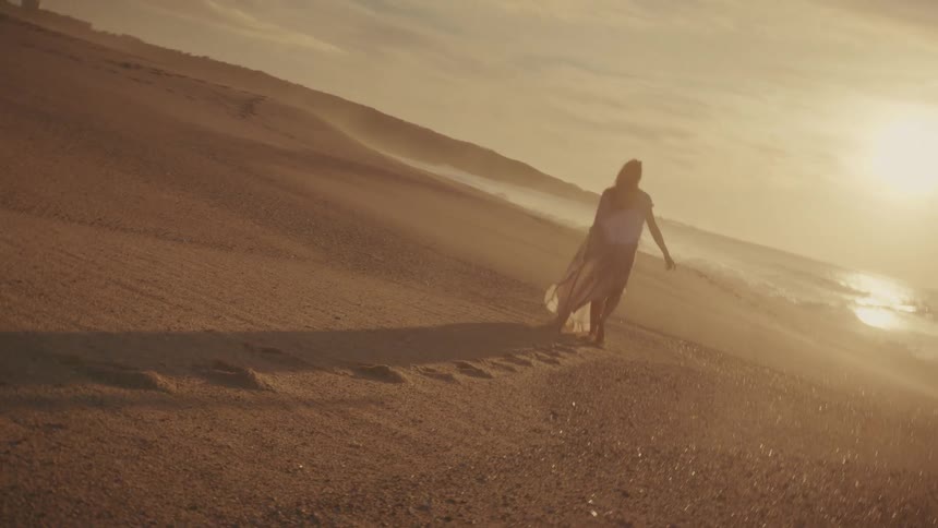 A woman walks along the beach at sunrise with her arms in the air