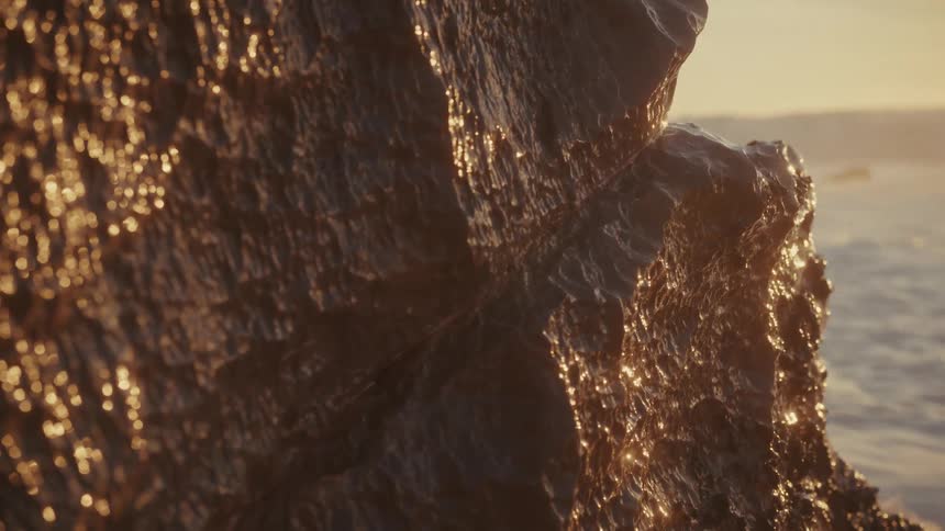 A closeup shot of a dark wet rock face on a beach intensely lit by a golden hour sun as a large ocean wave crashes and sprays white water across the foreground
