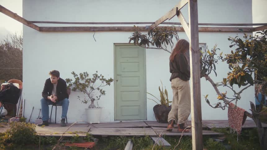 A young man sits on a chair while a young woman stands up from a crouch on the wooden porch of a small white house with a green door surrounded by an overgrown yard
