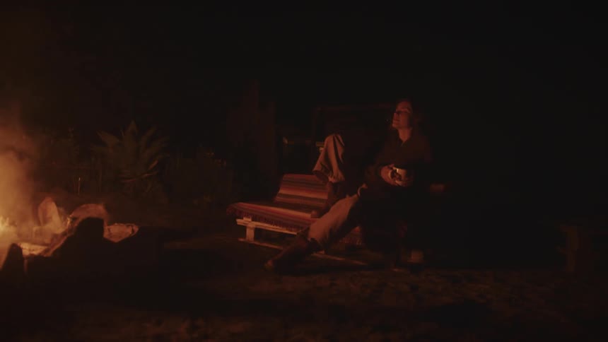 Woman sitting on a rustic bench watches a lit campfire at night
