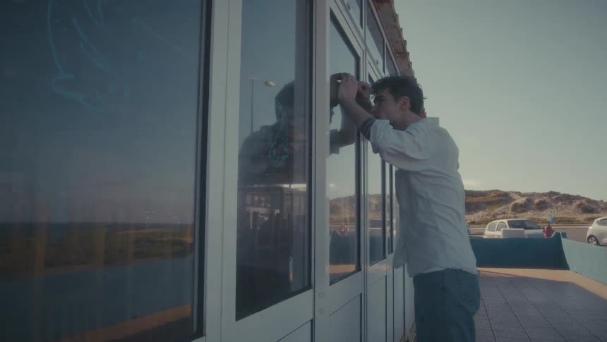 A young man in a lightcolored buttonup shirt and jeans leans his face against a large glass window peering intently inside a building there is a beach and mountains in the background