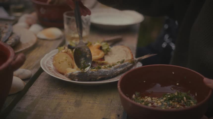 Person uses a spoon to season a sauce over baked fish with vegetables on a plate set on a rustic table