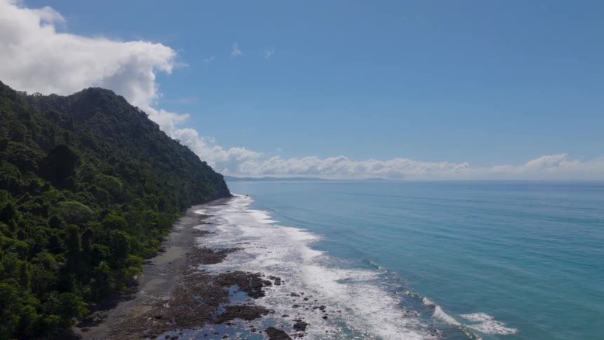 Green mountains lining rocky coast facing blue sea and sky with scattered clouds