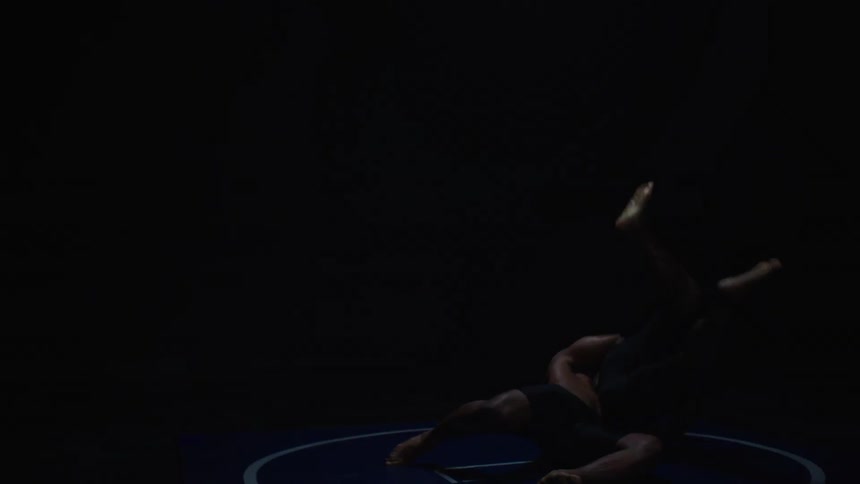Two shirtless men struggle in a dark room until one executes a takedown on the other bringing both wrestlers to the blue mat