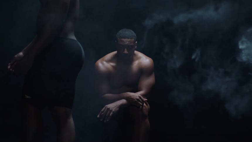 A figure walks past the camera to reveal a shirtless man sitting contemplatively in a dark smoky void