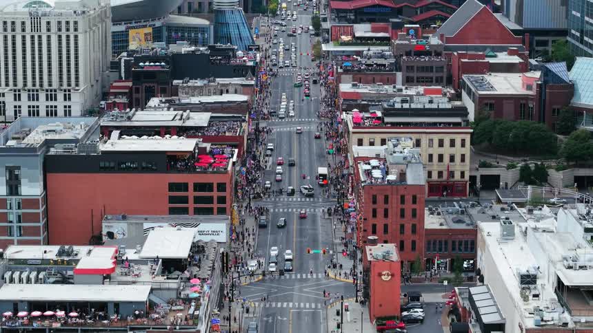 Pedestrians fill the sidewalks and cross the streets as traffic moves between downtown buildings with people on rooftops