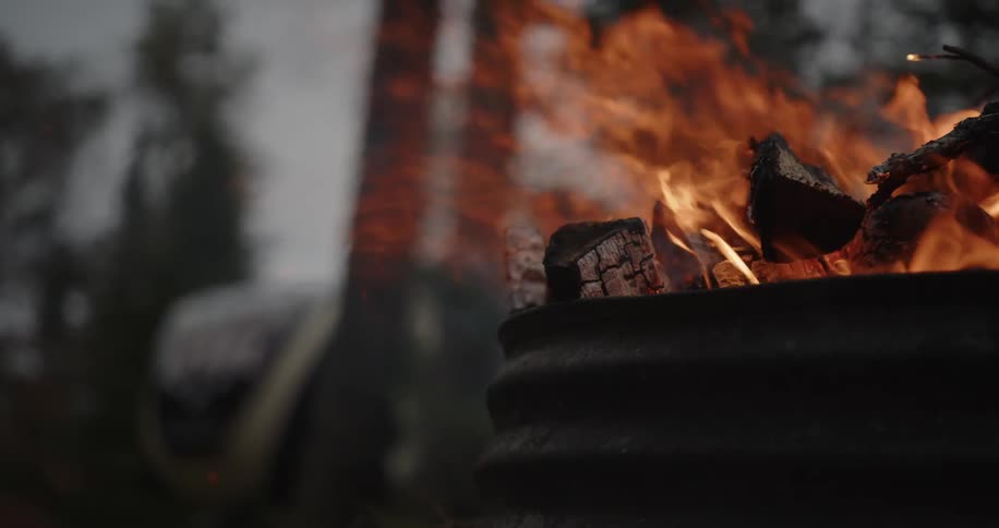 A close view of bright orange flames and smoke rising from burning wood logs in a dark metal fire pit outdoors in a hazy forest setting
