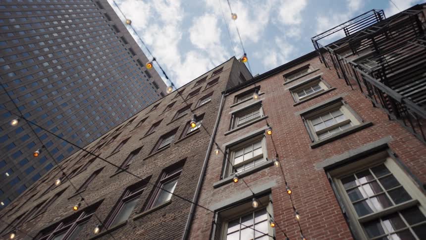From a low angle the camera slowly moves through a city alley looking up at brick buildings and a modern skyscraper as strings of lights hang overhead against a cloudy sky