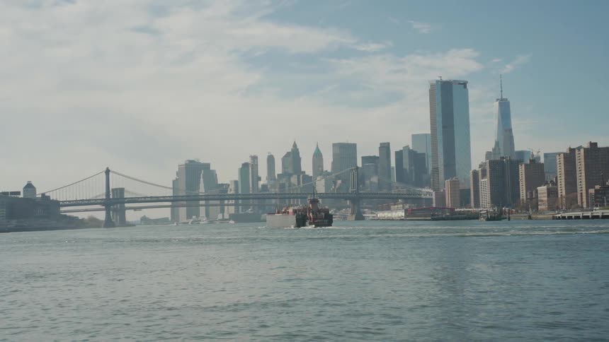 A wide long shot captures a city skyline and a suspension bridge across the water where a tugboat is seen moving away from the camera