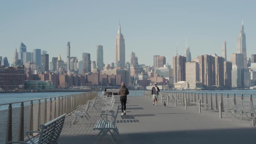 Man with a dog and other people enjoy a boardwalk across the water from the slightly foggy city during the daytime
