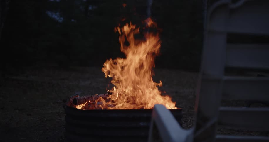 Wood burns inside a firepit outdoors with an empty chair in front of it at evening time