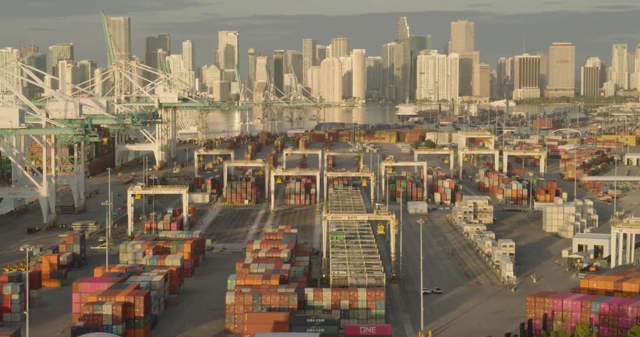 The scene is an impressive aerial view of a massive busy shipping terminal filled with stacks of colorful container boxes heavy machinery and rows of vehicle carriersin the background the modern skyline of a major city rises dramatically across the
