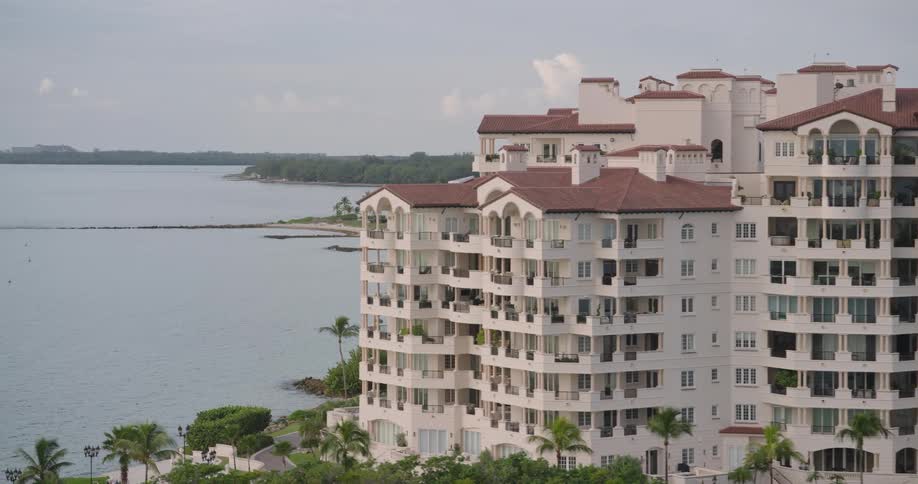 A slow panning shot reveals multistory white luxury residential buildings with red tiled roofs lining a manicured tropical beach protected by a rock seawall against the wide grayblue coastal water