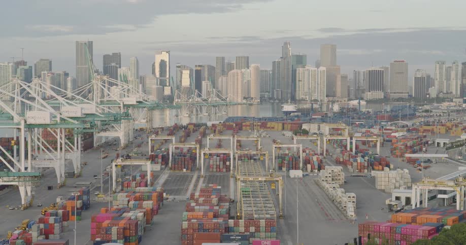 An aerial camera slowly pulls back to reveal the immense scale of a bustling port where cranes and trucks move colorful shipping containers against a modern city skyline