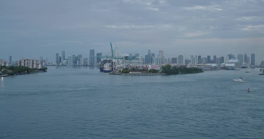 The video shows a wide view of a modern cityscape with many highrise buildings across a body of water under a cloudy skyin the foreground a port facility with large container cranes and docked ships is visible on a small landmass with boats moving i