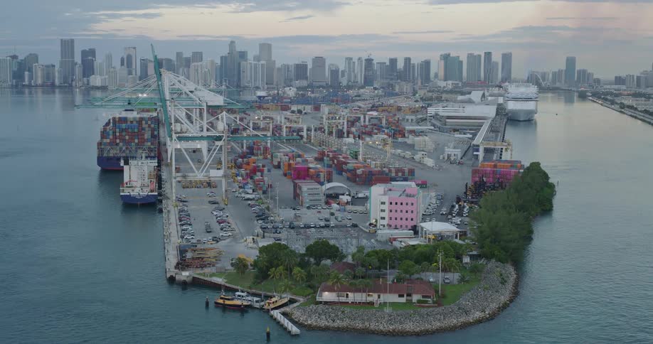 An aerial view shows a port with ships and cranes with a distant cityscape skyline visible
