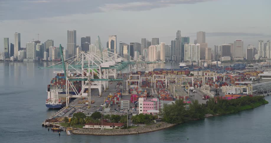 An elevated panoramic view captures a vast commercial shipping port with enormous white and green cranes and stacks of containers set against the backdrop of a dense modern city skyline by the water