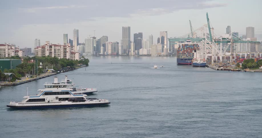 An elevated wide shot captures two white ferries moving along a channel in the foreground with residential buildings to the left and a dense city skyline and commercial shipping port in the distance