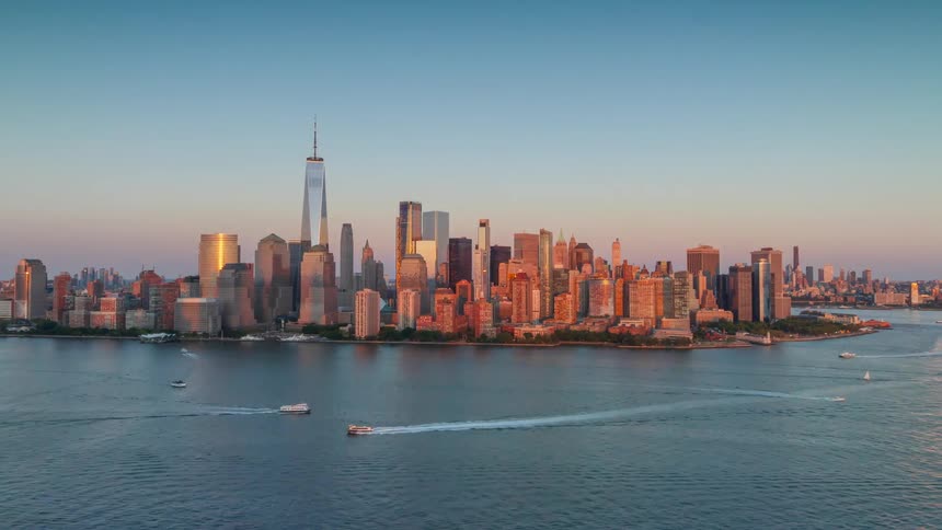 An aerial view of the new york city skyline at sunset with tall skyscrapers reflecting warm golden light above the calm water of the bay where several small boats are moving