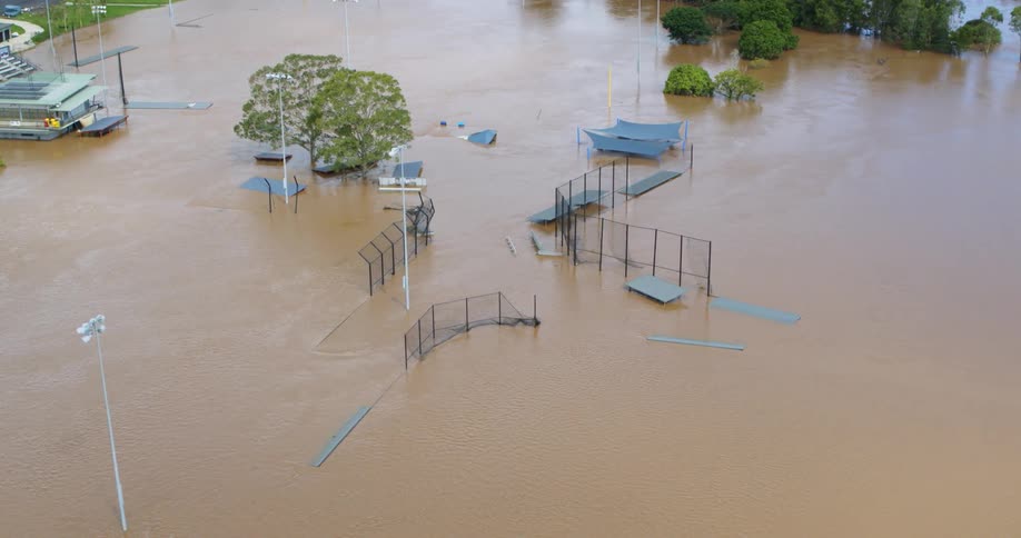 An aerial shot captures a large open recreational area severely flooded with fastmoving muddy brown water structures like fences ramps and shade coverings are partially submerged highlighting the extent of the flood damage