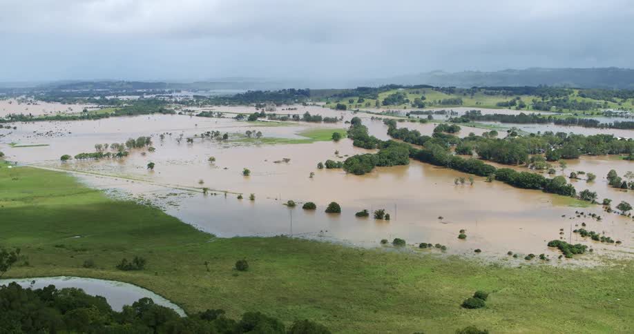 An aerial view captures widespread flooding across a rural landscape where brown waters completely submerge the fields and surround isolated clusters of trees beneath a somber grey sky