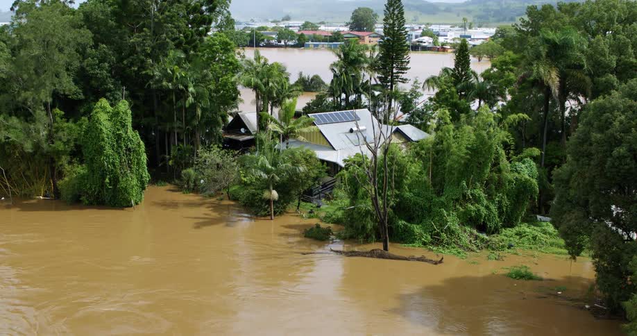House surrounded by vegetation appears partially flooded by muddy waters near an overflowing river