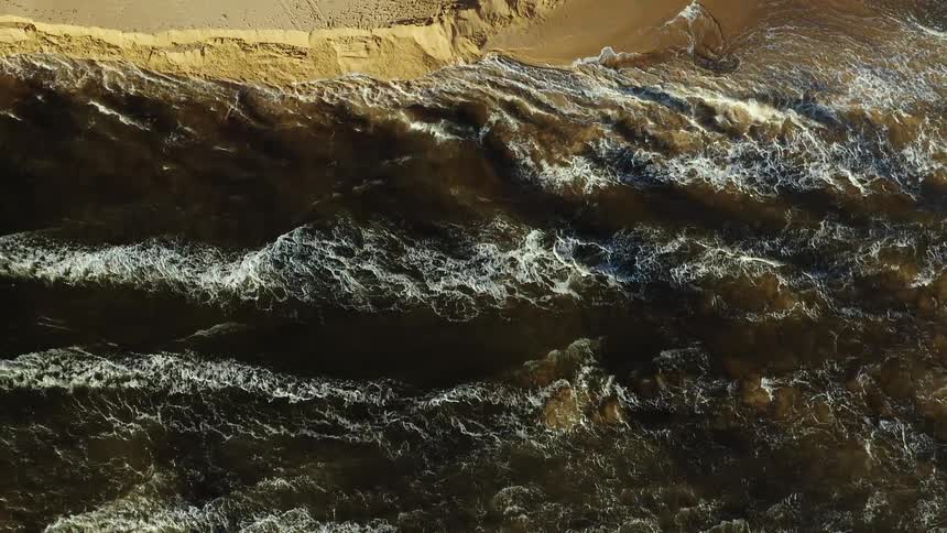 Aerial view of a dark churning waves crashing against a sandy shoreline