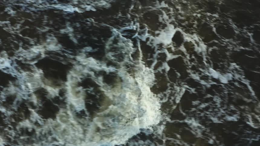 An aerial view shows dark powerful river rapids churning violently creating large patches of white foam and turbulence on the deep brown water