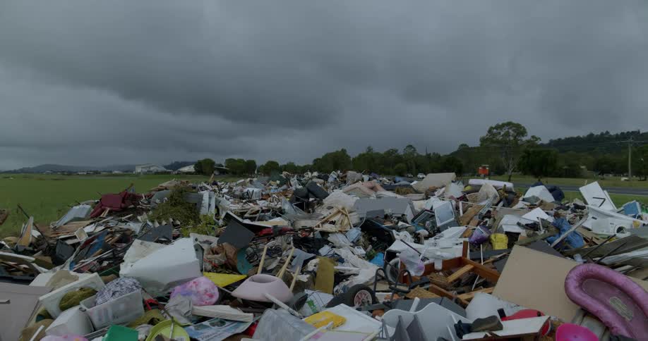 Trash including chairs and other household objects lie in a field near a highway on an overcast day