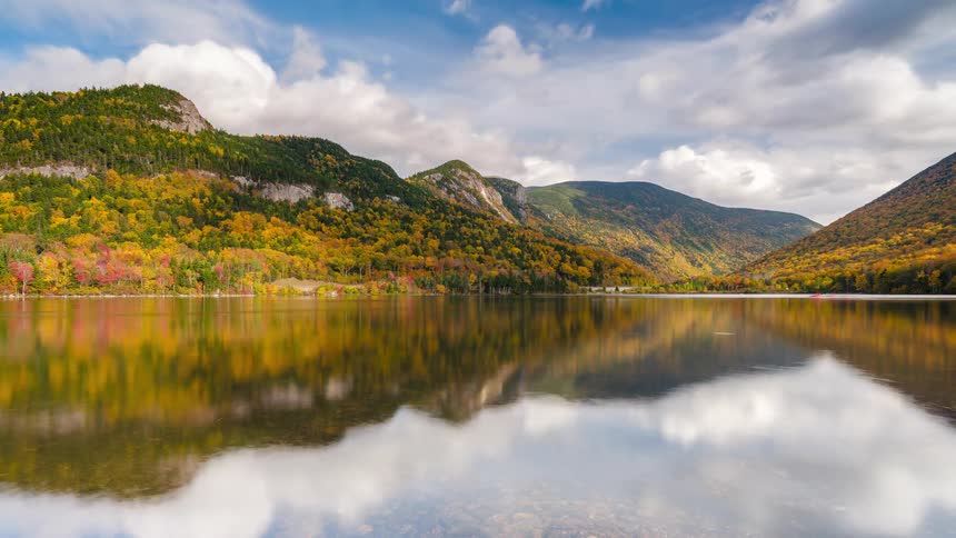 The tranquil lake with boaters show the dramatic rugged peaks of the surrounding mountains which are densely covered in vibrant autumn foliage under a bright sky dotted with soft white clouds