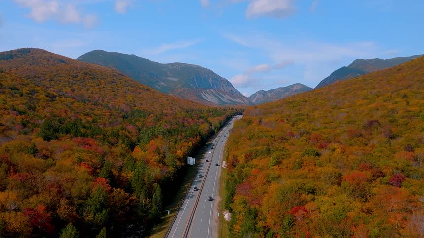 An aerial view follows a highway winding through a mountain valley surrounded by a dense forest ablaze with vibrant autumn colors