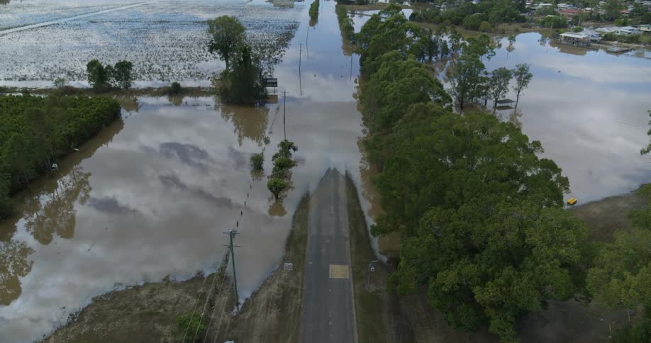 An aerial view captures a road partially submerged by extensive murky brown floodwaters across a flat landscape the still water reflects the overcast sky and the line of trees highlighting the impact of the flooding on the community