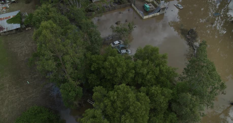 An overhead drone shot captures severe flooding in a neighborhood showing murky brown water covering the streets and yards with several cars partially submerged among the trees and houses