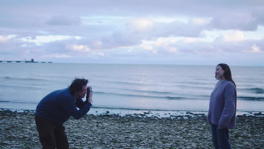 Person being photographed on rocky beach facing ocean under cloudy sky
