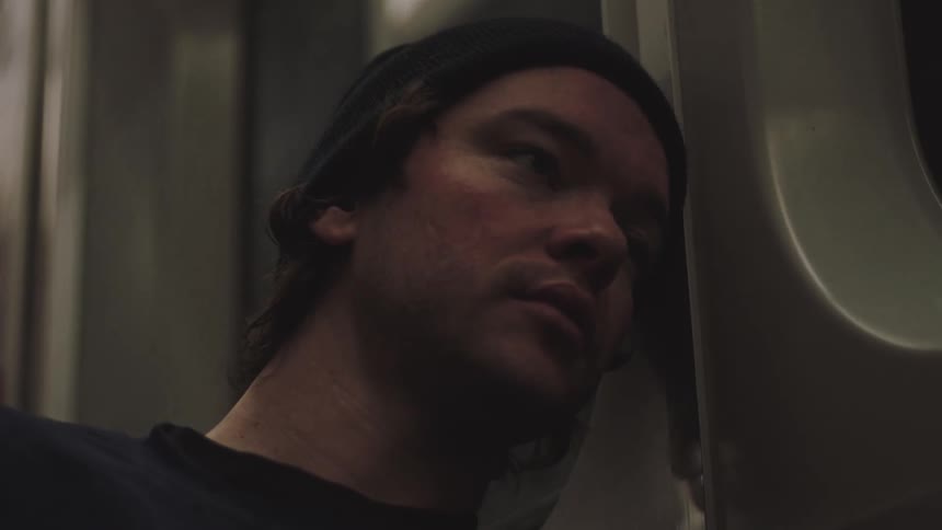Young adult man wearing a black beanie leaning his head against the window of a train indoors