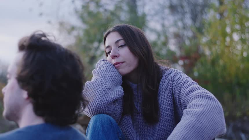 A woman sits thoughtfully outdoors watching her boyfriend off camera she speaks to him briefly as he sits down beside her