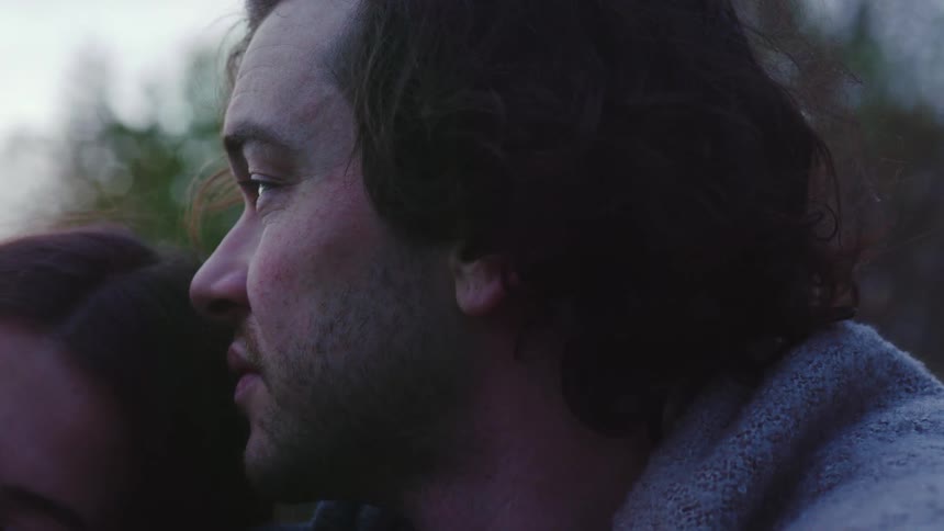 Young adult man talking to a young adult woman while she rests her head on his chest outdoors during dusk or dawn