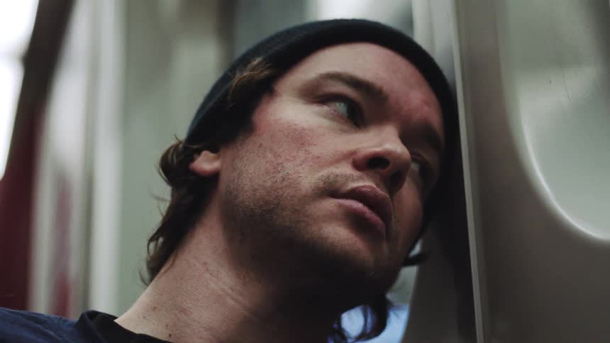 A closeup of a man with a beanie sleeping while leaning against the curved wall of a moving subway who briefly opens his eyes before resting again