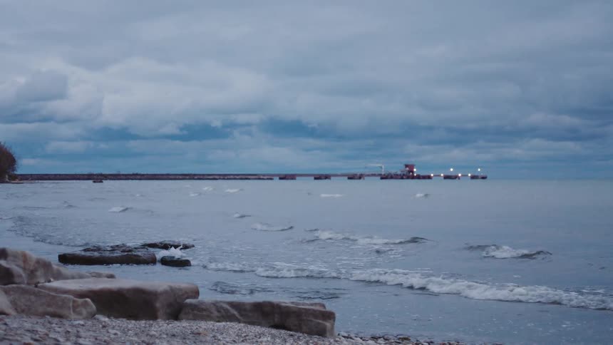 Bluegrey waves roll gently onto a rocky shoreline washing past large stones in the foreground while a distant pier glows faintly under the heavy overcast sky