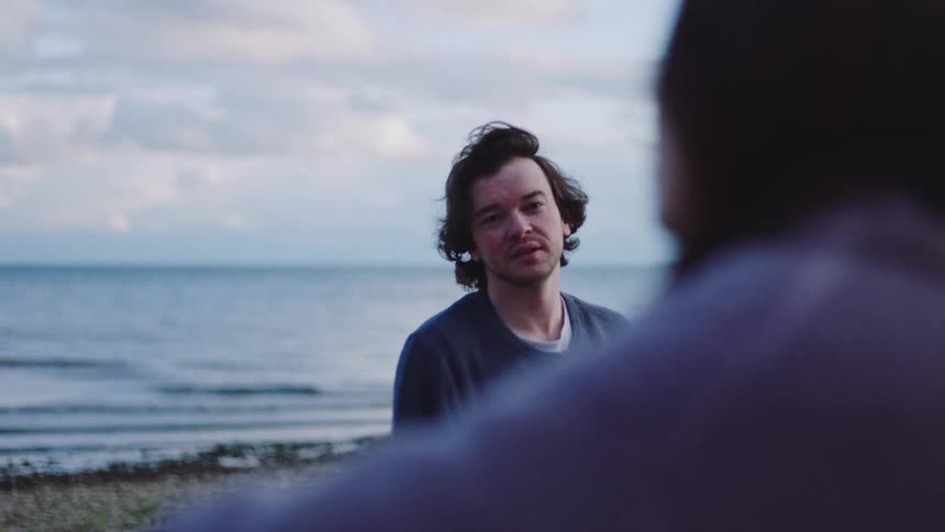 A young man standing on a beach with the ocean and sky in the background makes a throwing motion before turning and smiling at a person sitting out of focus in the foreground then turns his attention back to the water