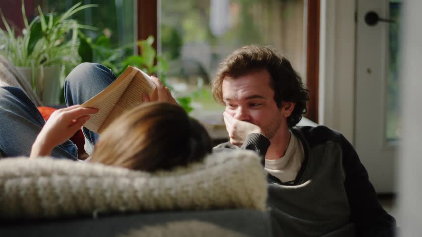 Couple reclining on a sofa near a window while reading books in a naturally lit environment
