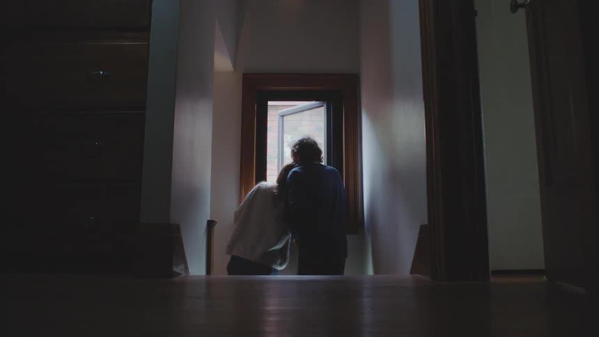 Framed in a dark interior two people stand at an open doorway embracing tightly in a moment of emotional farewell with their backs to the camera
