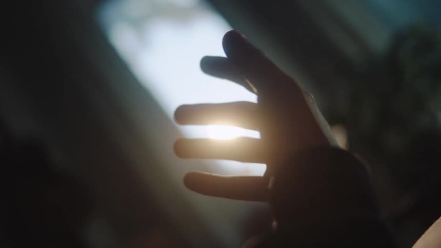 A close view of a dark silhouetted hand slowly reaching up toward a bright white light source creating a moody atmosphere