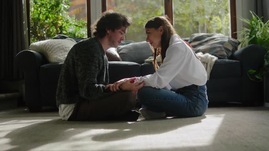 A man and a woman sit crosslegged on a living room floor holding hands and leaning close for an intimate and supportive moment bathed in natural light from a large window